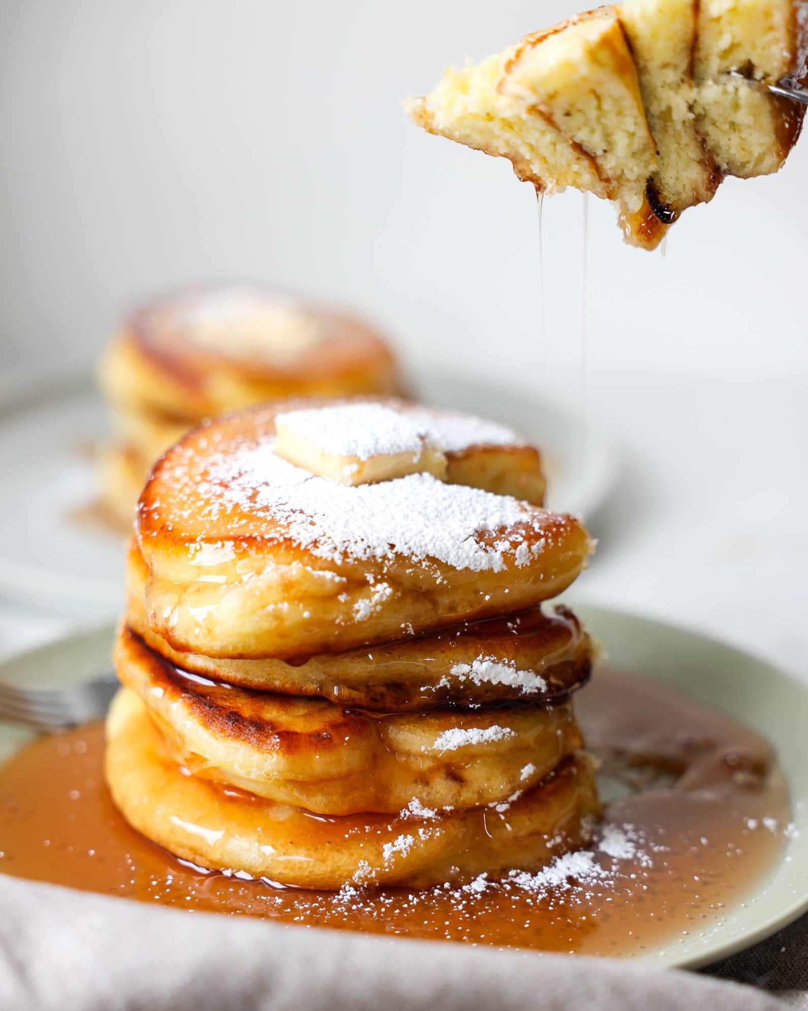 Stack of pancakes with syrup and powdered sugar on a plate, with a piece being lifted.
